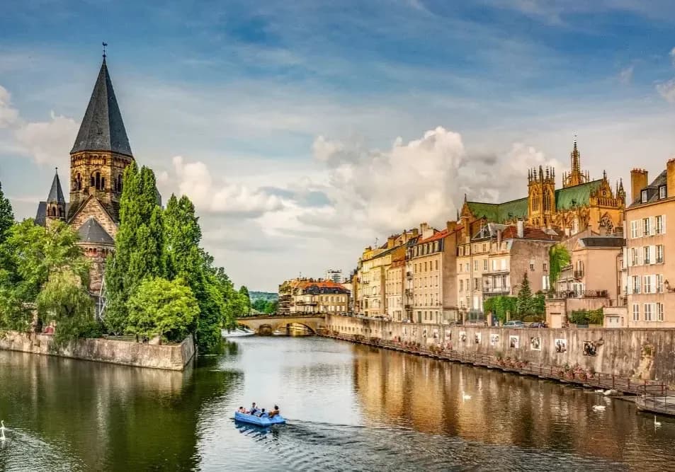 Vue panoramique de Metz avec le Temple Neuf au bord de la Moselle, architecture historique et bâtiments colorés longeant la rivière sous un ciel bleu.