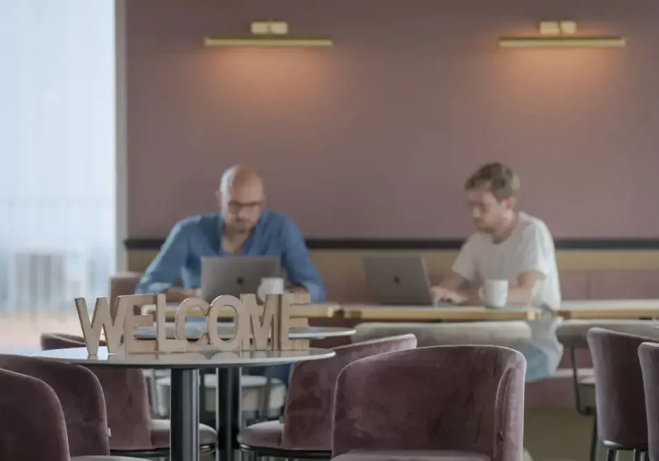 Espace de coworking chaleureux avec signe "WELCOME" en bois au premier plan et deux professionnels concentrés travaillant en arrière-plan.
