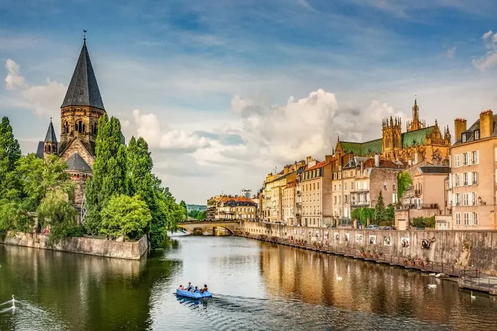 Vue panoramique de Metz avec le Temple Neuf au bord de la Moselle, architecture historique et bâtiments colorés longeant la rivière sous un ciel bleu.