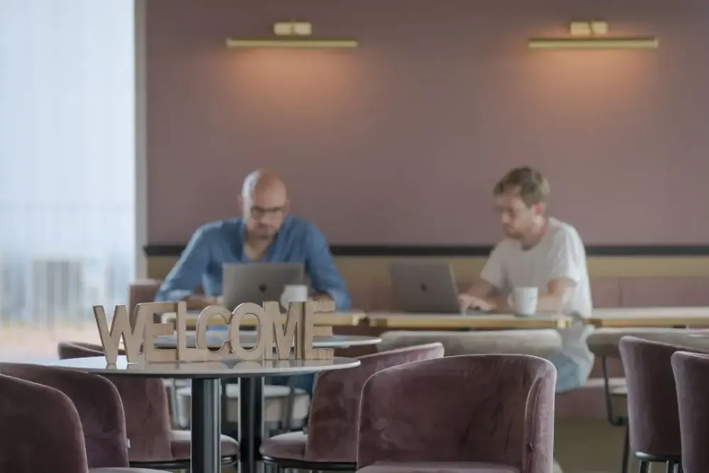 Espace de coworking chaleureux avec signe "WELCOME" en bois au premier plan et deux professionnels concentrés travaillant en arrière-plan.