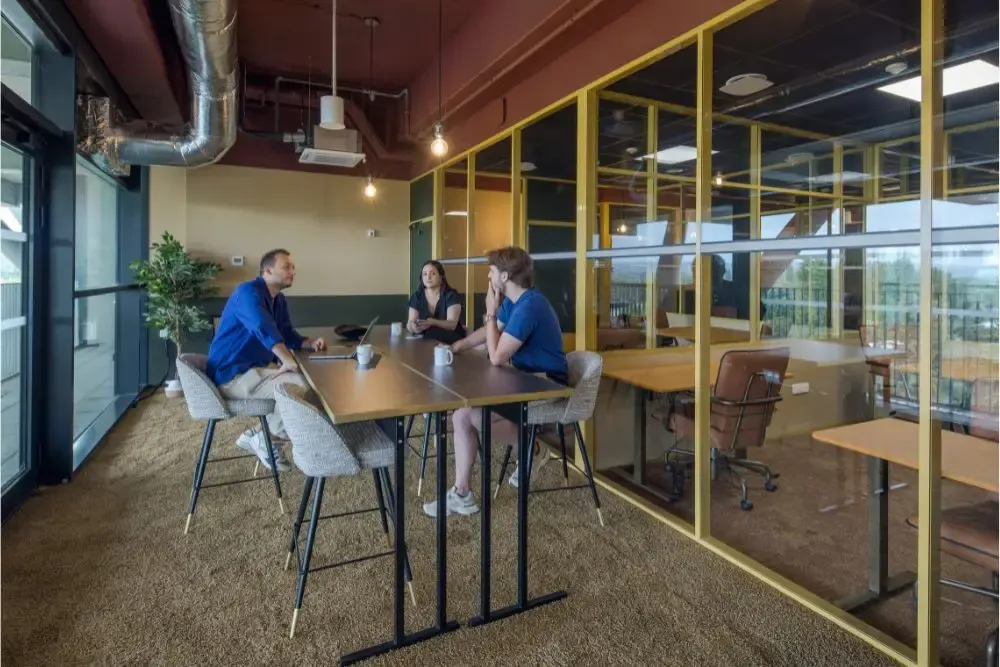 Trois professionnels discutant autour d'une table haute dans un espace de coworking à Metz, avec grandes baies vitrées et espaces de travail privatifs visibles.