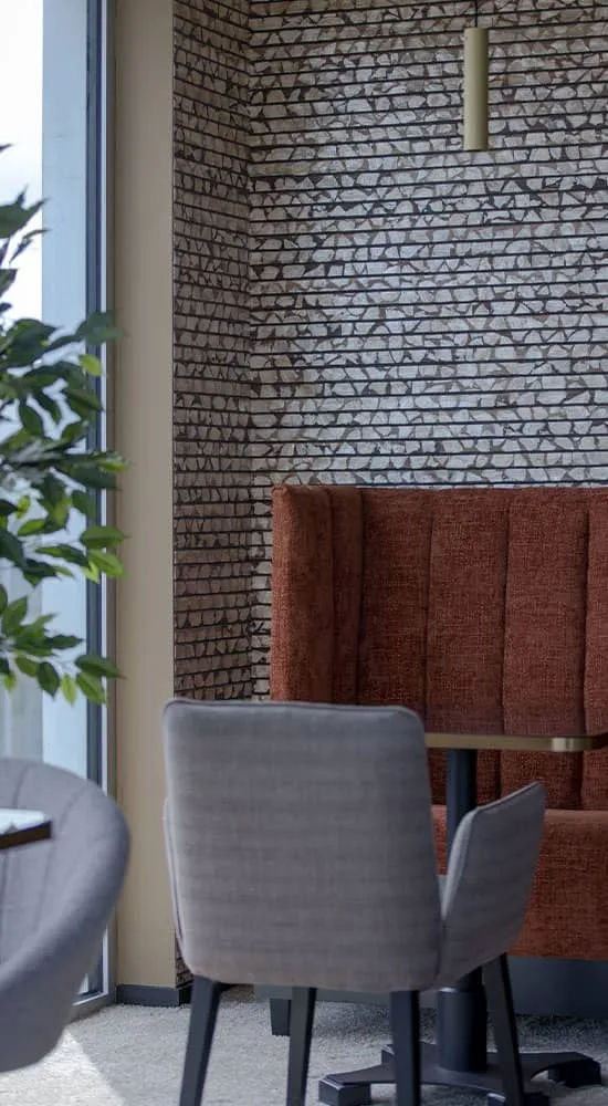 Une alcôve de coworking chaleureuse avec un canapé en tissu brun contre un mur en briques décoratives, flanqué de chaises modernes en tissu gris, le tout éclairé par la lumière naturelle qui se déverse de la fenêtre adjacente, incarnant l'alliance de confort et de design chez NOW Coworking Nantes.