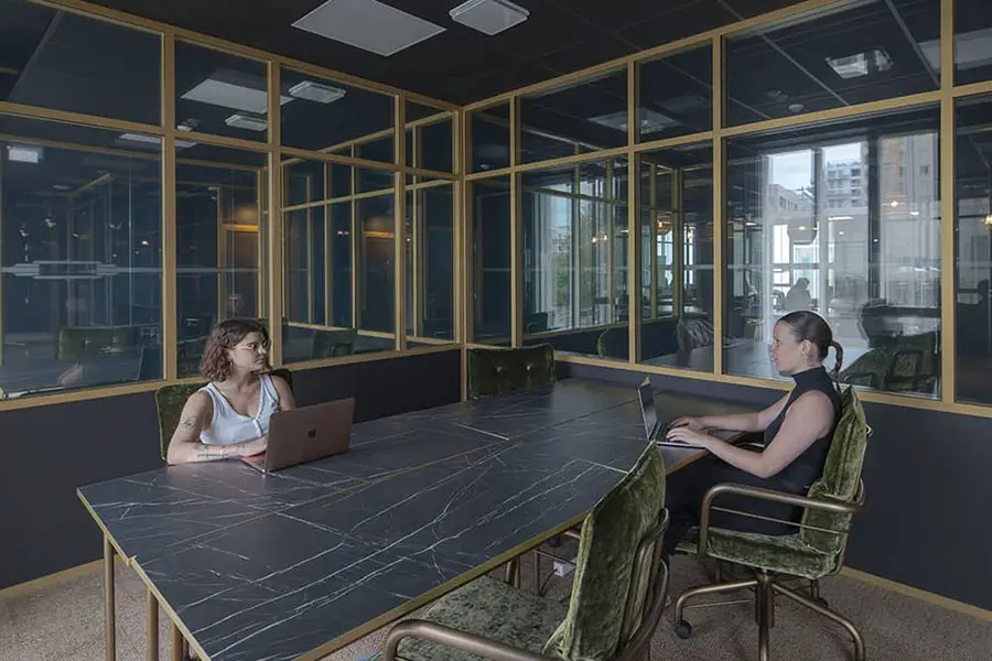 Deux professionnels travaillant en face à face dans un bureau partagé à Nantes, avec des tables en marbre noir et des chaises en velours vert, entourés de cloisons vitrées avec des cadres dorés, alliant intimité et transparence.