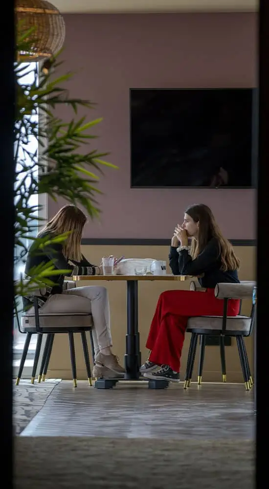 Vue à travers une ouverture sur deux collègues prenant une pause dans un espace de coworking à Nantes, assises autour d'une table haute, dans un cadre paisible avec une décoration intérieure élégante et des touches de verdure.